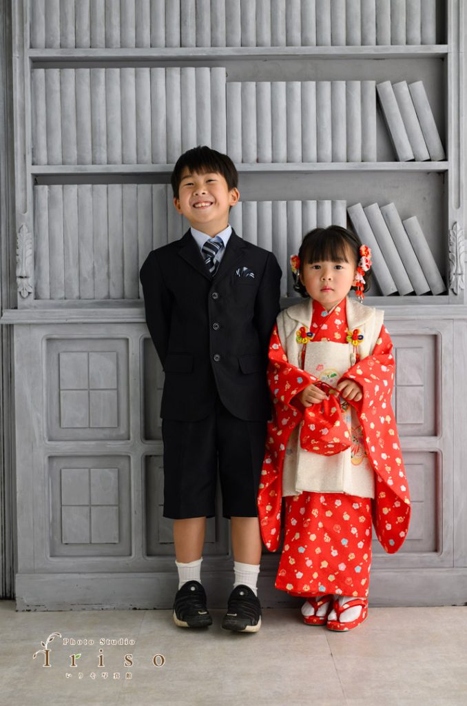 黒いスーツ姿の男の子と赤い着物を着た女の子が本棚の前で並んで立つ微笑ましい姿。
