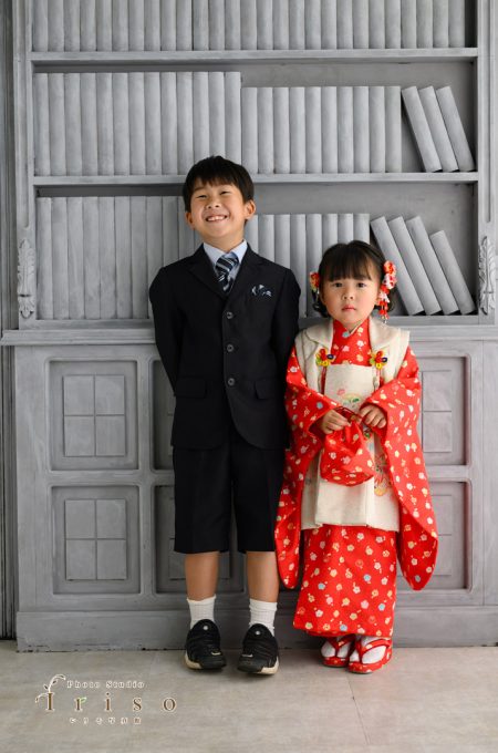 黒いスーツ姿の男の子と赤い着物を着た女の子が本棚の前で並んで立つ微笑ましい姿。