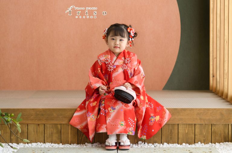 鮮やかな赤い着物を着た女の子が、伝統的な座敷に座り穏やかな表情で前を見つめている姿。