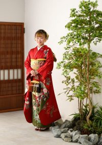 赤色の華やかな振袖、緑の葉が茂る木の前で笑顔を見せている。振袖には花や草の模様が施され、帯は金色を基調にしたデザイン。少女は伝統的な和装バッグを手に持ち、背景には和風の引き戸が見える。