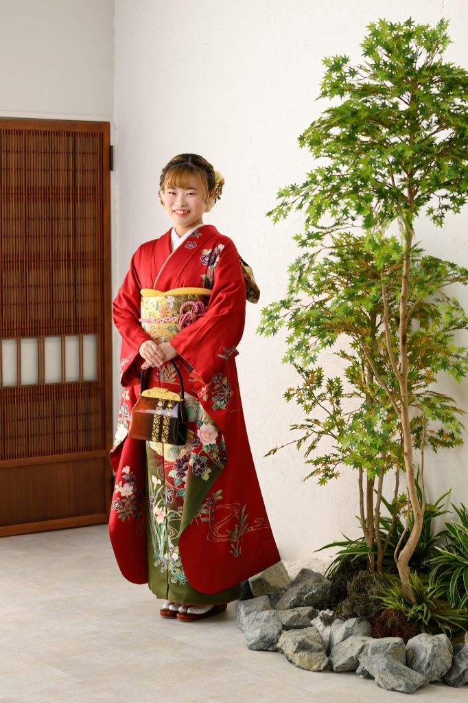 赤色の華やかな振袖、緑の葉が茂る木の前で笑顔を見せている。振袖には花や草の模様が施され、帯は金色を基調にしたデザイン。少女は伝統的な和装バッグを手に持ち、背景には和風の引き戸が見える。