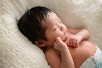 新生児が柔らかな毛布の上で眠っている愛らしい姿。小さな手を握り、穏やかで安心した表情が印象的。淡い自然光が赤ちゃんの肌を優しく照らし、温かく穏やかな雰囲気が漂う一枚。
