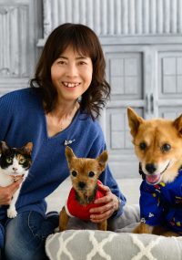 青いセーターを着た女性が、三毛猫と2匹の犬と一緒に座っている。女性は笑顔でカメラを見つめ、右側の犬は青い服を着てクッションの上に座っている。