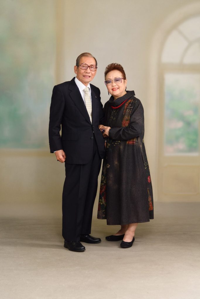 フォーマルな服装をした高齢の男性と女性が、手を取り合って微笑んでいる。男性は黒いスーツとネクタイ、女性は黒を基調とした柄のあるドレスを着用し、眼鏡をかけている。背景には窓があり、穏やかな雰囲気が漂う室内での撮影。