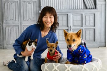 青いセーターを着た女性が、三毛猫と2匹の犬と一緒に座っている。女性は笑顔でカメラを見つめ、右側の犬は青い服を着てクッションの上に座っている。
