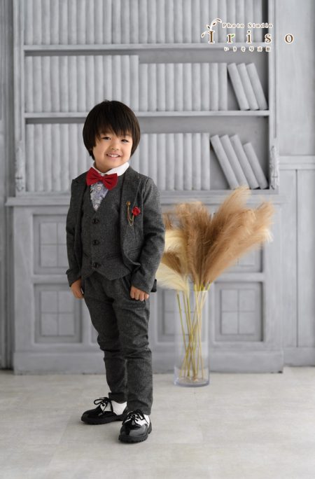 グレーのスーツに赤い蝶ネクタイを合わせた男の子が笑顔で立ち、背景に本棚とパンパスグラスがある写真。