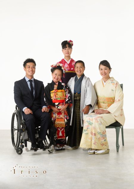 家族5人がフォーマルな衣装で並び、笑顔で記念撮影。父親は車椅子に座り、母親と子供たちは和装で優雅にポーズ。