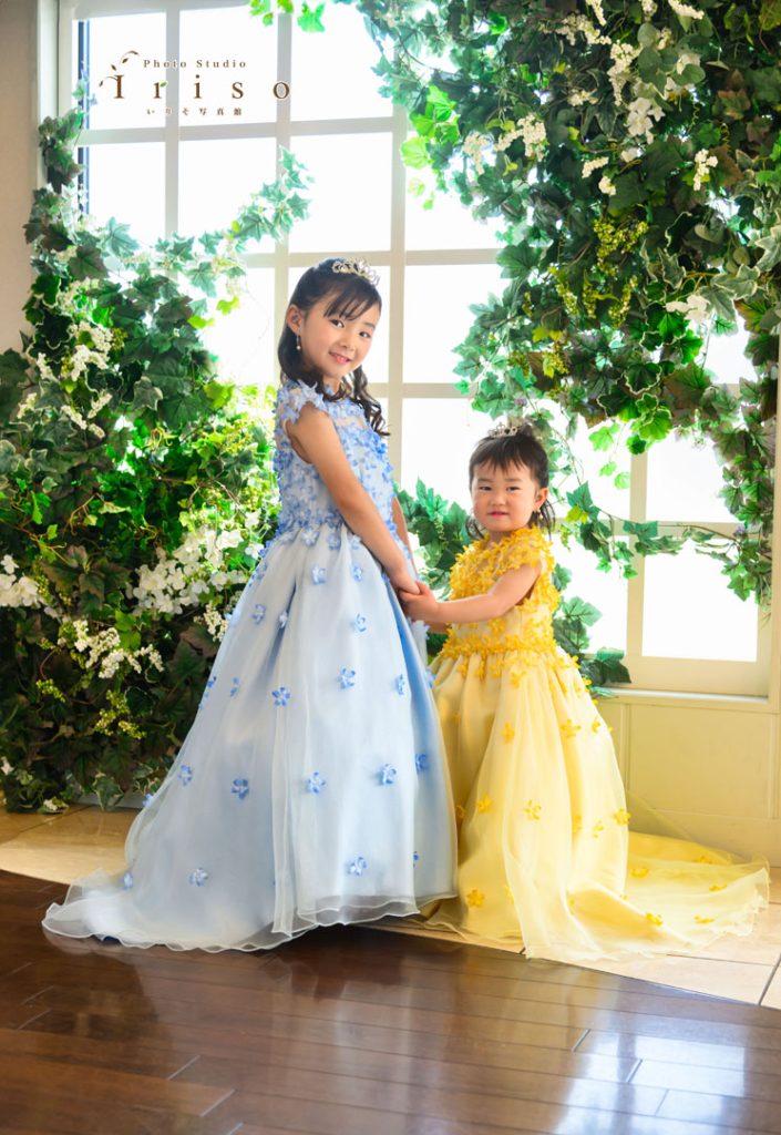 青と黄色のフラワードレスを着た姉妹が、笑顔で手をつなぎ緑の装飾がある窓辺でポーズをとる。
