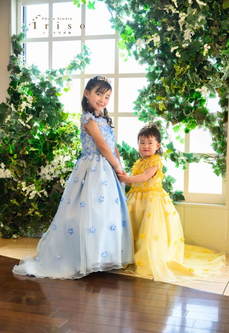 青と黄色のフラワードレスを着た姉妹が、笑顔で手をつなぎ緑の装飾がある窓辺でポーズをとる。