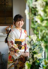 白地に赤と黒の花柄振袖を着た女性が、窓辺で微笑みながら立つ写真。周囲には緑の装飾が施されています。