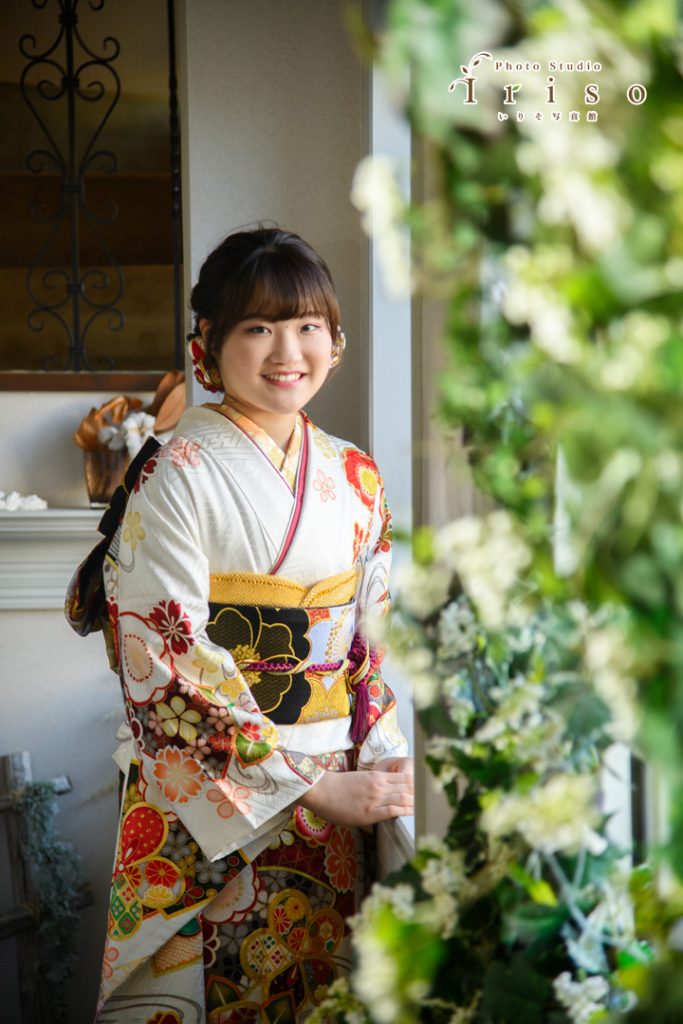 白地に赤と黒の花柄振袖を着た女性が、窓辺で微笑みながら立つ写真。周囲には緑の装飾が施されています。