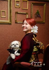 深紅の着物を着た女性が和装姿の小型犬を抱え、微笑みながら椅子に座る姿。