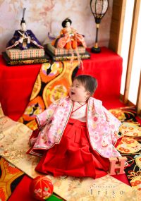ひな人形の飾りの前で、赤い袴とピンクの着物を着た赤ちゃんが笑顔を見せている写真。