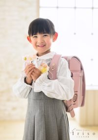 ピンクのランドセルを背負い、手に花を持った笑顔の女の子が、新入学を祝う姿で撮影されています。