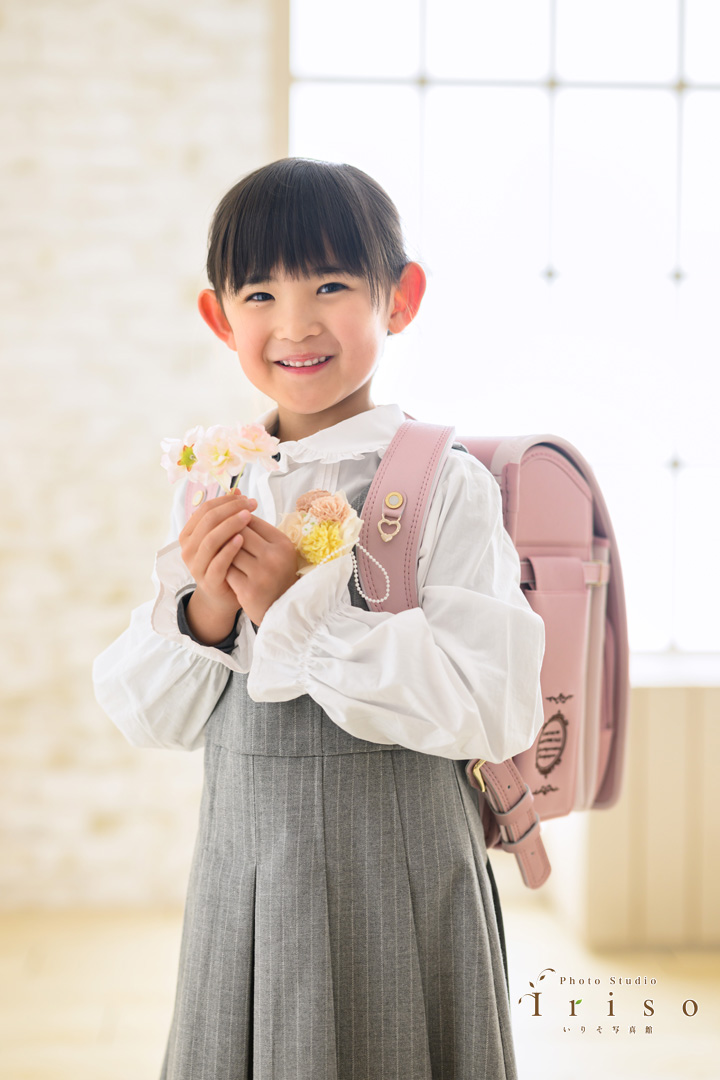 ピンクのランドセルを背負い、手に花を持った笑顔の女の子が、新入学を祝う姿で撮影されています。