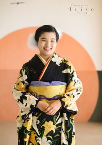 黒地に白い花柄の振袖を着た女性が、鮮やかな黄色の帯を締めて笑顔で立っている写真。