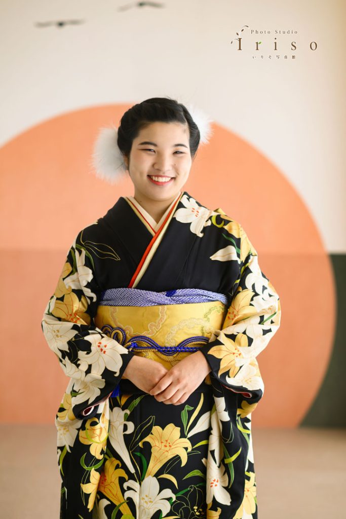 黒地に白い花柄の振袖を着た女性が、鮮やかな黄色の帯を締めて笑顔で立っている写真。