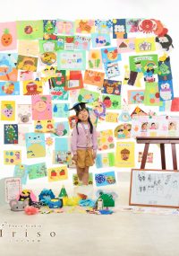 手作りの絵や工作が壁一面に飾られた前で、メダルを首にかけた笑顔の女の子が立っています。