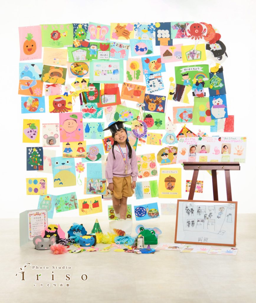 手作りの絵や工作が壁一面に飾られた前で、メダルを首にかけた笑顔の女の子が立っています。