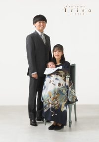 スーツ姿の父親と椅子に座った母親が、祝い着をまとった赤ちゃんを抱えながら撮影された家族写真。