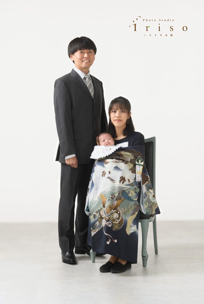 スーツ姿の父親と椅子に座った母親が、祝い着をまとった赤ちゃんを抱えながら撮影された家族写真。