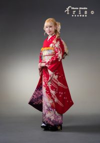 赤い花柄の振袖を着た女性が、金と紫の帯を締めて上品にポーズを取る姿。