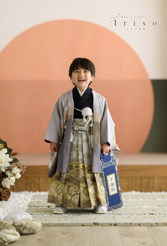 金とグレーの華やかな袴を着た男の子が、千歳飴の袋を手に持ちながら笑顔で立っています。