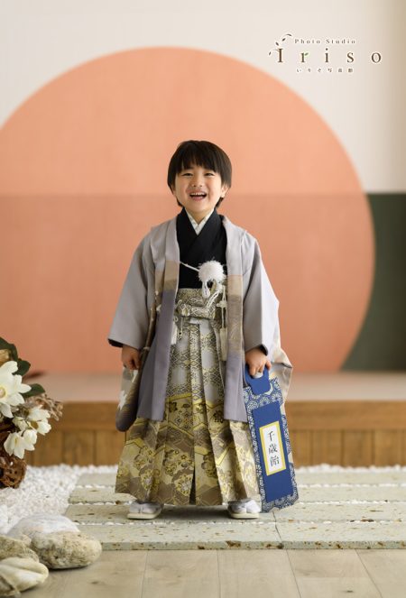 金とグレーの華やかな袴を着た男の子が、千歳飴の袋を手に持ちながら笑顔で立っています。