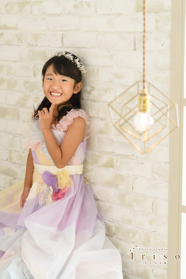 パステルカラーのフリル付きドレスを着た笑顔の女の子が、白いレンガの壁にもたれてポーズをとっている様子を描いています。前景には、装飾的な幾何学模様のペンダントライトが温かみのある雰囲気