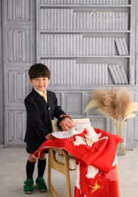 スーツ姿の男の子が、赤い祝い着に包まれた赤ちゃんの頭に優しく手を添えながら笑顔を見せています。
