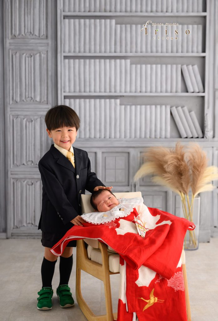 スーツ姿の男の子が、赤い祝い着に包まれた赤ちゃんの頭に優しく手を添えながら笑顔を見せています。