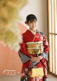 赤と黒の花柄振袖を着た女性が、金色の帯とバッグを持ち、微笑みながら立つ写真。