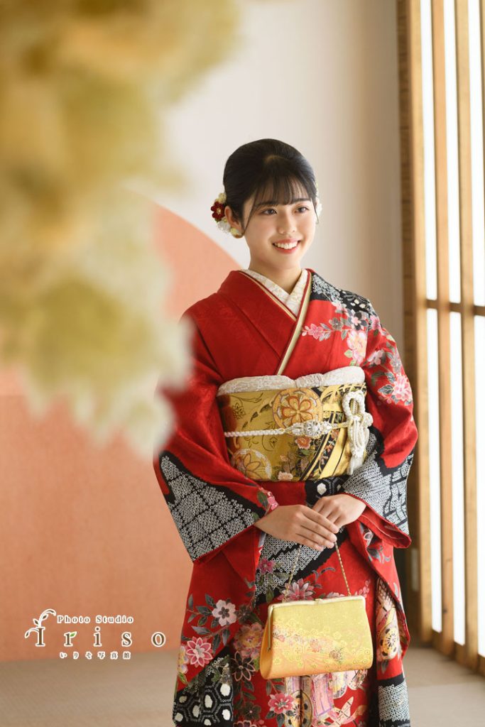 赤と黒の花柄振袖を着た女性が、金色の帯とバッグを持ち、微笑みながら立つ写真。