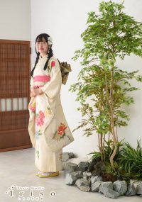 花柄の白い振袖を着た女性が、日本庭園風の飾りのそばで微笑みながら立つ姿。