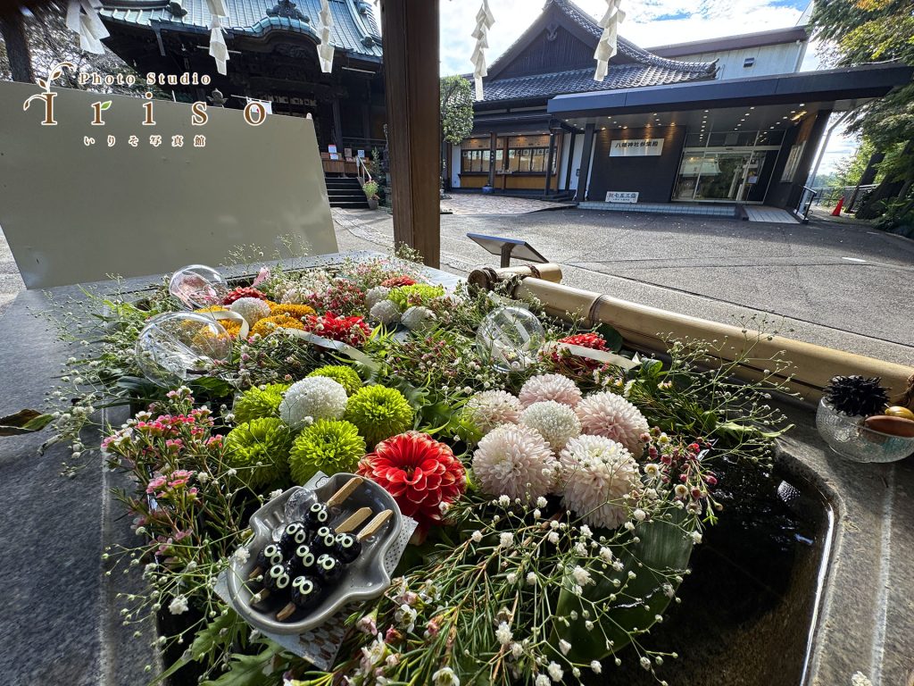 狭山の神社境内に美しく飾られた花々と、背景には伝統的な建物が広がる風景。おみくじや花装飾が和の趣を演出