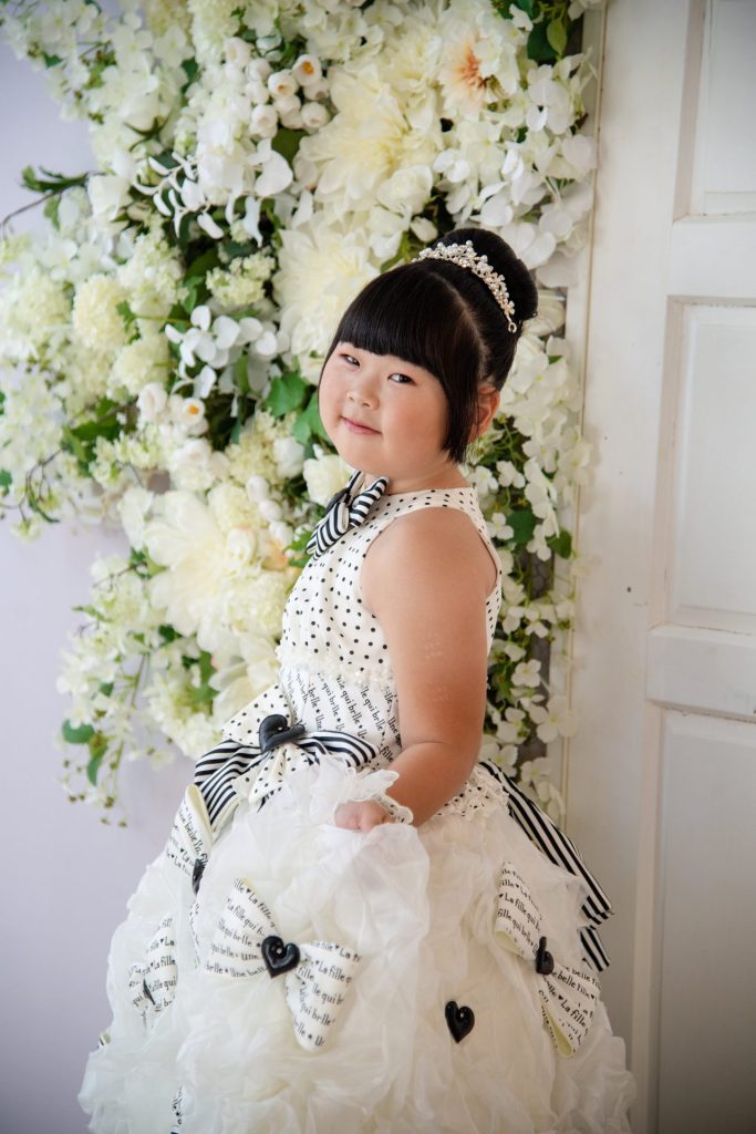 純白のドレスを着て、白い花の壁の前で微笑む女の子の写真