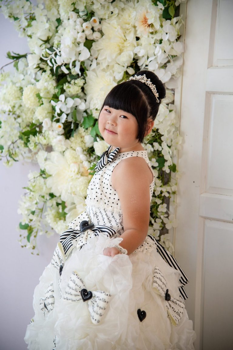純白のドレスを着て、白い花の壁の前で微笑む女の子の写真