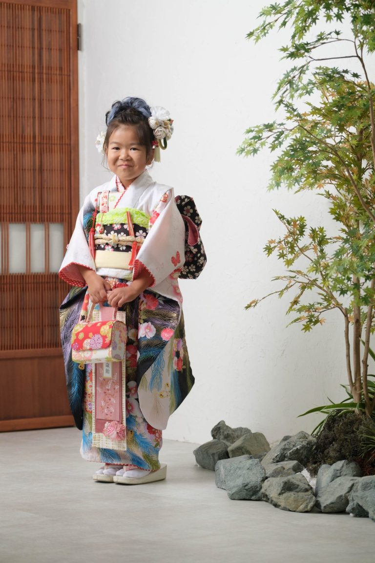 七五三の着物姿の女の子が、少しはにかんだ笑顔でカメラを見ている写真。華やかな着物に身を包んだお嬢様の、なんとも愛らしい笑顔！少し緊張している様子もまた可愛いですね。