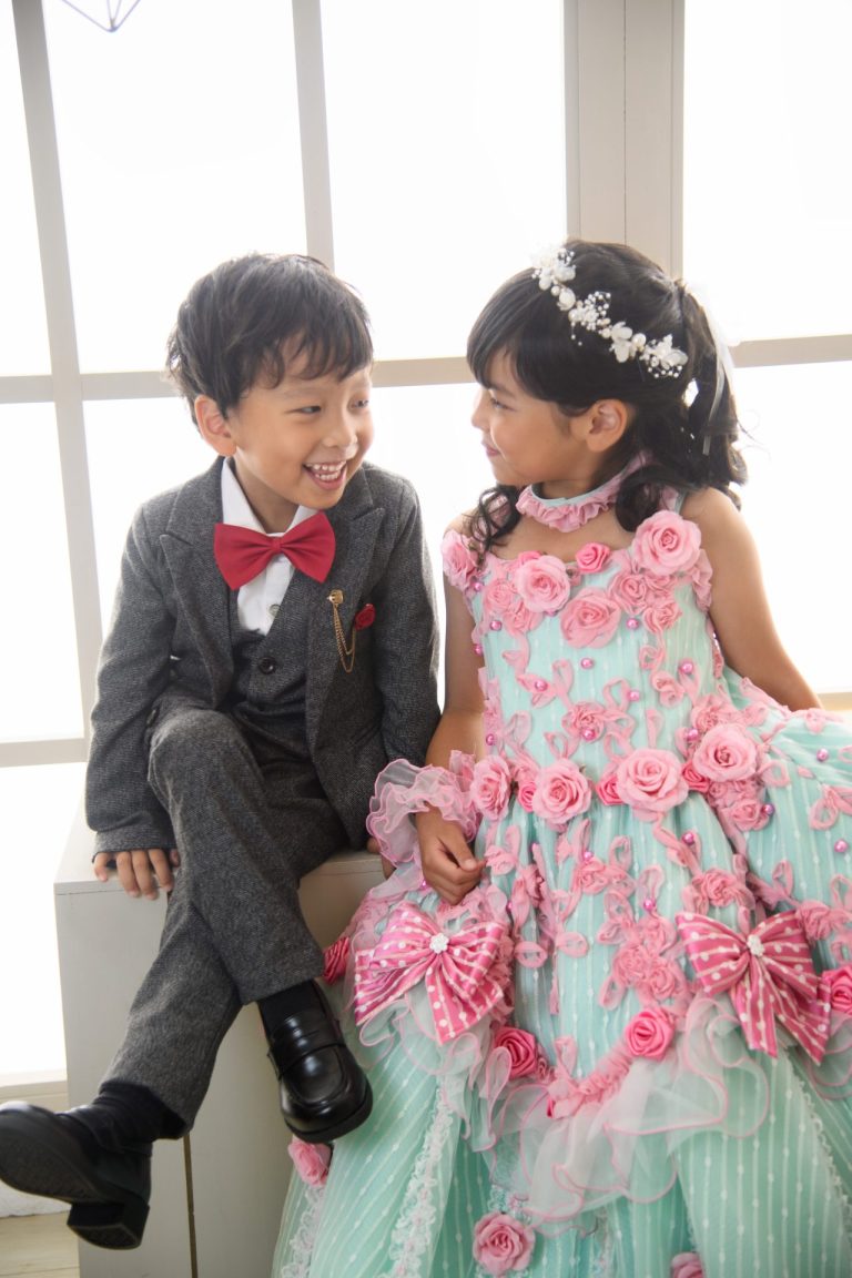窓辺でドレス姿の女の子とスーツ姿の男の子が並んで座り、笑顔で話している様子。 淡いミントグリーンのドレスにピンクの薔薇が散りばめられたドレス姿の女の子と、 グレーのスーツに赤い蝶ネクタイが映える男の子。