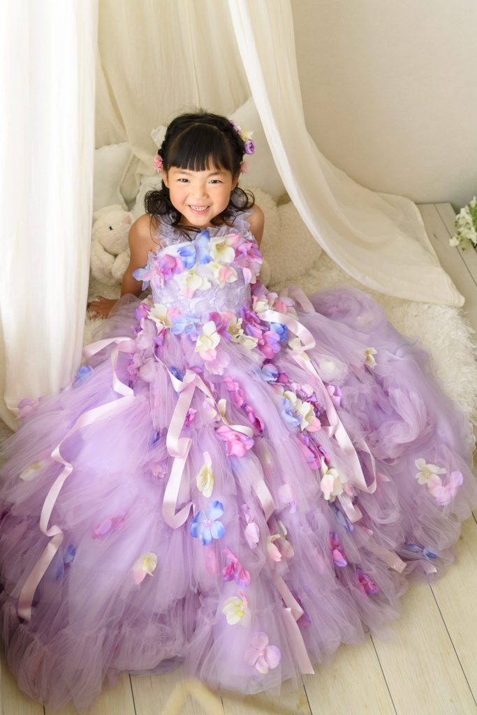 パステルパープルのドレスが、まるで満開のお花畑みたい！カラフルなお花とリボンが散りばめられたドレスは、女の子の可愛らしさをより一層引き立てています。ふんわりとしたチュールスカートも、夢のような雰囲気。