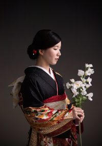 着物姿で白い花束を持った女性の横顔写真。黒地に金糸の刺繍が施された振袖に、華やかな帯を合わせた素敵なコーディネートですね。白い花束が清楚な雰囲気をさらに引き立てています。