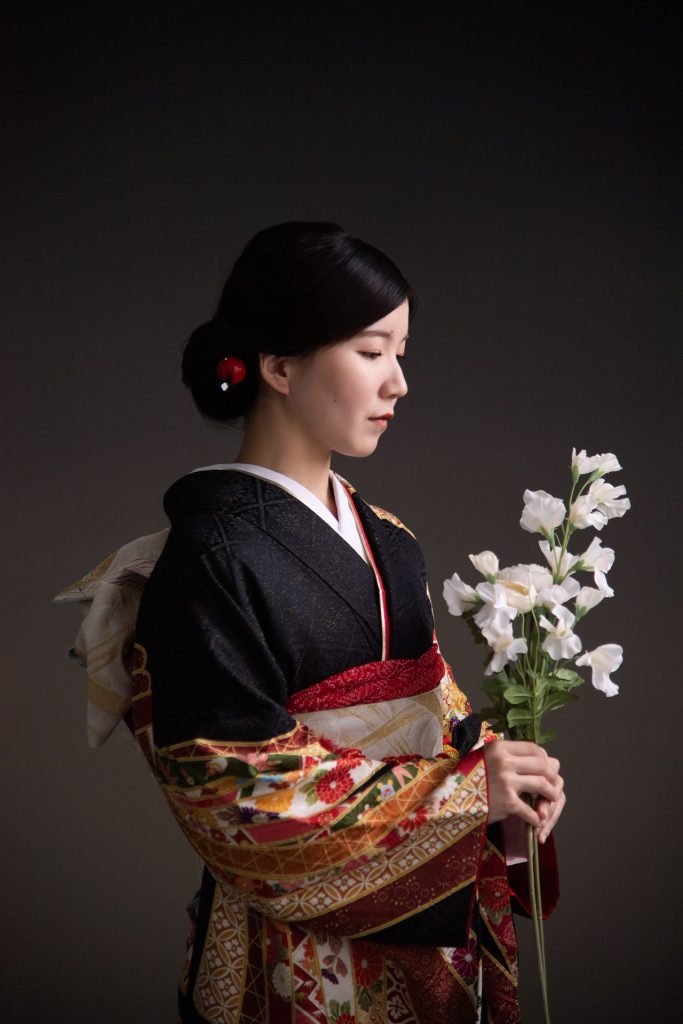 着物姿で白い花束を持った女性の横顔写真。黒地に金糸の刺繍が施された振袖に、華やかな帯を合わせた素敵なコーディネートですね。白い花束が清楚な雰囲気をさらに引き立てています。