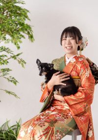 振袖姿の女性が愛犬を抱っこして笑顔の写真。鮮やかなオレンジ色の振袖に身を包み、大切な愛犬を優しく抱きかかえる姿が印象的です。満面の笑みからは、成人式を迎えられた喜びと、愛犬への愛情が溢れ出ていますね。
