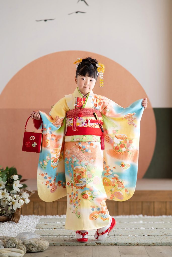 七五三の着物を着て、少し照れながらも嬉しそうに微笑む女の子の写真。淡い黄色の着物に、赤い帯が映える、華やかな装い。