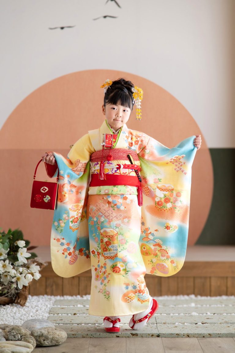 七五三の着物を着て、少し照れながらも嬉しそうに微笑む女の子の写真。淡い黄色の着物に、赤い帯が映える、華やかな装い。