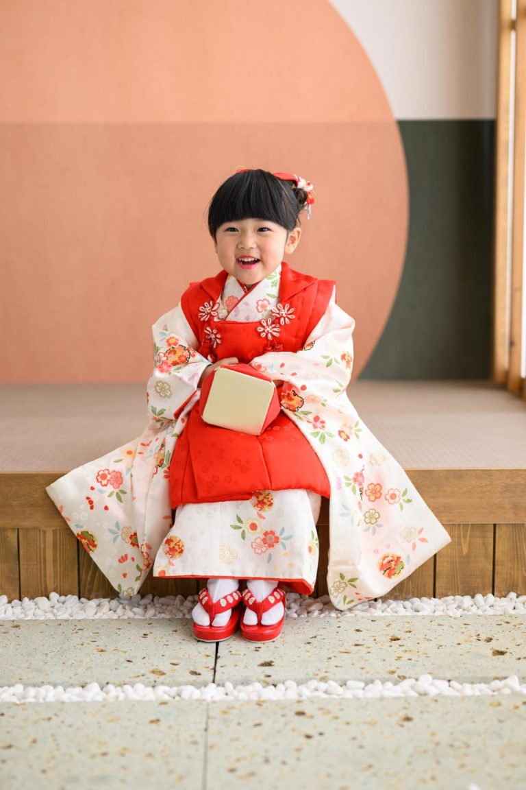 七五三衣装を着た女の子が、少し緊張した面持ちでちょこんと座っている様子。 赤い被布と白い着物がよくお似合いです！ちょっぴり緊張しているのかな？という表情。でも、ふとした瞬間に見せる笑顔がとっても可愛らしかったです。