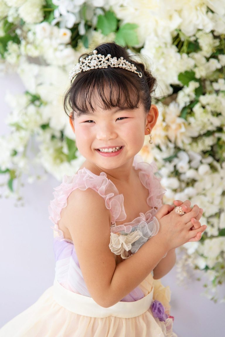 ドレスを着てティアラをつけた女の子の笑顔の写真。満開の白い花々に囲まれて、まるで物語のお姫様のような姿！淡いパステルカラーのドレスとキラキラ輝くティアラが、彼女の可愛らしさを一層引き立てています。