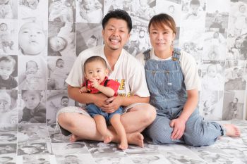 これまでの成長を撮りためてきて数々の写真をモノクロで並べて、その写真の前で１歳近年の家族写真